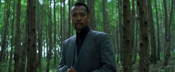 Movie still from “Predators” (2010), directed by Nimród Antal – A man standing in a wooded area wearing a suit and tie; Close Up shot, Low angle