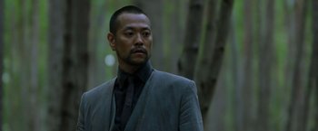 Movie still from “Predators” (2010), directed by Nimród Antal – A person wearing a suit near a tree; Close Up shot, Low angle