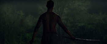 Movie still from “Predators” (2010), directed by Nimród Antal – A shirtless man holding a sword in the dark; Medium shot, Over the shoulder angle
