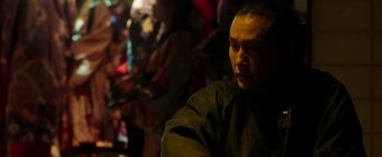 Movie still from “Prisoners of the Ghostland” (2021), directed by Sion Sono – A man sitting in front of a group of people; Close Up shot, Over the shoulder angle