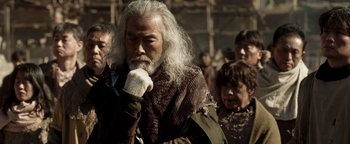 Movie still from “Prisoners of the Ghostland” (2021), directed by Sion Sono – An old man with long white hair and a beard; Medium shot, Over the shoulder angle