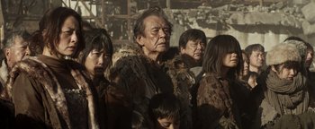 Movie still from “Prisoners of the Ghostland” (2021), directed by Sion Sono – An old man in a group of people; Medium shot, Over the shoulder angle