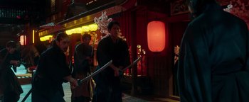 Movie still from “Prisoners of the Ghostland” (2021), directed by Sion Sono – A man holding a pair of swords while standing in front of a building; Medium shot, Low angle