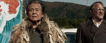 Movie still from “Prisoners of the Ghostland” (2021), directed by Sion Sono – An older man wearing a fur coat standing in front of a car; Medium shot, Low angle