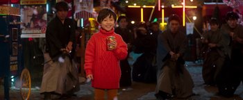 Movie still from “Prisoners of the Ghostland” (2021), directed by Sion Sono – A young boy in a red sweater is holding a box of candy; Medium shot, Over the shoulder angle