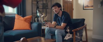 Movie still from “Puppy Love” (2023), directed by Richard Alan Reid – A man sitting on a couch holding a cell phone next to a dog; Medium shot, Over the shoulder angle