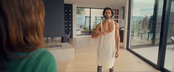 Movie still from “Puppy Love” (2023), directed by Richard Alan Reid – A man in a white tank top is standing in a living room; Medium shot, Over the shoulder angle