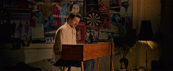 Movie still from “Puppy Love” (2023), directed by Richard Alan Reid – A man sitting in front of a piano in front of a dartboard; Wide shot, High angle