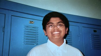 Movie still from “Queenmaker: The Making of an It Girl” (2023), directed by Zackary Drucker – A young man wearing glasses standing in front of two blue lockers; Extreme Close Up shot, Overhead angle