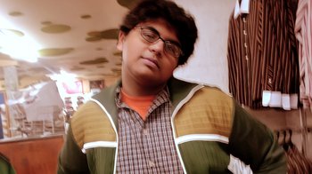 Movie still from “Queenmaker: The Making of an It Girl” (2023), directed by Zackary Drucker – A young man with glasses and a brown and green jacket; Close Up shot, Low angle