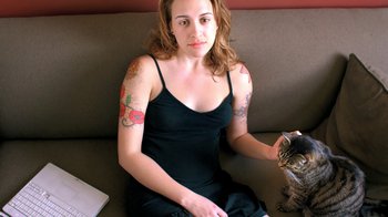 Movie still from “Queenmaker: The Making of an It Girl” (2023), directed by Zackary Drucker – A woman sitting on a couch holding a cat; Close Up shot, High angle