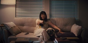 Movie still from “Quiz Lady” (2023), directed by Jessica Yu – A woman sitting on a couch holding a remote control; Medium shot, Over the shoulder angle