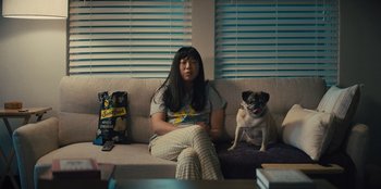 Movie still from “Quiz Lady” (2023), directed by Jessica Yu – A woman sitting on a couch next to a pug dog; Medium shot, Over the shoulder angle