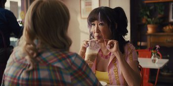 Movie still from “Quiz Lady” (2023), directed by Jessica Yu – A woman with purple hair is drinking from a cup; Medium shot, Over the shoulder angle