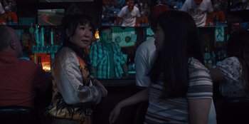 Movie still from “Quiz Lady” (2023), directed by Jessica Yu – Two women sitting in front of a television in front of a crowd; Medium shot, Over the shoulder angle