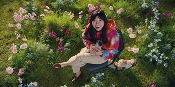 Movie still from “Quiz Lady” (2023), directed by Jessica Yu – A woman sitting on a chair holding a camera; Wide shot, High angle