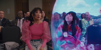 Movie still from “Quiz Lady” (2023), directed by Jessica Yu – A group of people sitting in front of an image of a girl; Medium shot, Over the shoulder angle