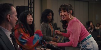 Movie still from “Quiz Lady” (2023), directed by Jessica Yu – A group of people sitting around a room; Medium shot, Over the shoulder angle