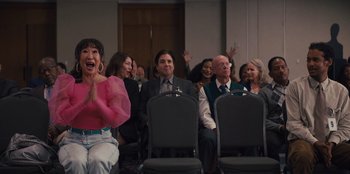 Movie still from “Quiz Lady” (2023), directed by Jessica Yu – A group of people sitting in a room with their hands in the air; Medium shot, Over the shoulder angle