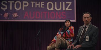 Movie still from “Quiz Lady” (2023), directed by Jessica Yu – A woman sitting in front of a microphone in front of an audience; Medium shot, Over the shoulder angle