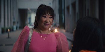 Movie still from “Quiz Lady” (2023), directed by Jessica Yu – A woman in a pink dress is smiling for the camera; Medium shot, Over the shoulder angle