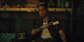 Movie still from “Quiz Lady” (2023), directed by Jessica Yu – A man sitting on a couch holding a hair brush; Medium shot, Over the shoulder angle