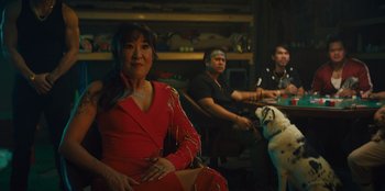 Movie still from “Quiz Lady” (2023), directed by Jessica Yu – A group of people sitting in a room with a dog; Medium shot, Over the shoulder angle