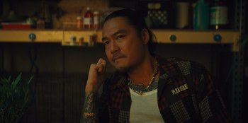 Movie still from “Quiz Lady” (2023), directed by Jessica Yu – A man with tattoos and a beard sitting in a room; Close Up shot, Low angle