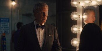 Movie still from “Quiz Lady” (2023), directed by Jessica Yu – A man in a suit standing in front of lights; Medium shot, Over the shoulder angle