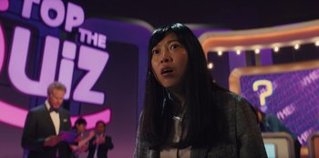 Movie still from “Quiz Lady” (2023), directed by Jessica Yu – A woman with long black hair standing in front of a microphone; Medium shot, Over the shoulder angle