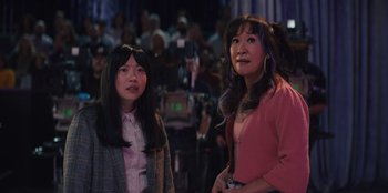Movie still from “Quiz Lady” (2023), directed by Jessica Yu – Two women standing next to each other in front of an audience; Medium shot, Over the shoulder angle