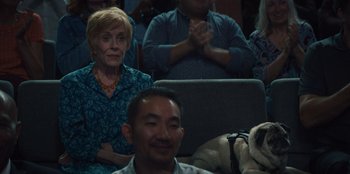 Movie still from “Quiz Lady” (2023), directed by Jessica Yu – A group of people sitting in front of a dog; Medium shot, Over the shoulder angle