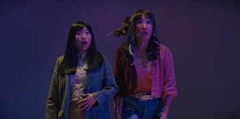 Movie still from “Quiz Lady” (2023), directed by Jessica Yu – Two women standing next to each other in front of a purple background; Medium shot, Low angle