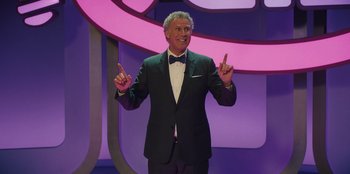 Movie still from “Quiz Lady” (2023), directed by Jessica Yu – A man in a suit and bow tie is giving the peace sign; Medium shot, Over the shoulder angle