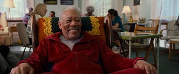 Movie still from “RED” (2010), directed by Robert Schwentke – An older man sitting in a chair smiling for the camera; Medium shot, Over the shoulder angle