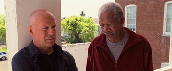 Movie still from “RED” (2010), directed by Robert Schwentke – Two men standing next to each other near a window; Medium shot, Low angle