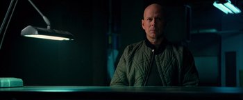 Movie still from “RED” (2010), directed by Robert Schwentke – A bald man sitting at a table in front of a microphone; Medium shot, Over the shoulder angle