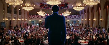 Movie still from “RED” (2010), directed by Robert Schwentke – A man standing in front of a crowd of people in a room; Wide shot, High angle