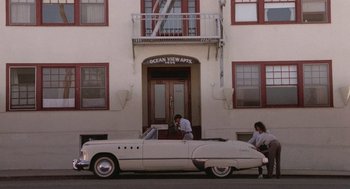 Movie still from “Rain Man” (1988), directed by Barry Levinson – Two people standing next to an old white car in front of a building; Extreme Wide shot, High angle