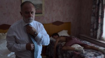 Movie still from “Rainbow” (2022), directed by Paco León – An older man adjusting his tie in front of a sleeping woman; Medium shot, Low angle