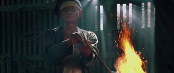 Movie still from “Rambo” (2008), directed by Sylvester Stallone – An old man is holding a stick over a fire; Medium shot, Low angle