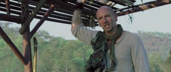 Movie still from “Rambo” (2008), directed by Sylvester Stallone – A bald man wearing a scarf and holding onto a wooden structure; Close Up shot, Low angle
