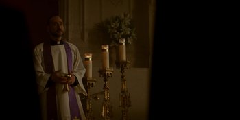 Movie still from “Ray Donovan: The Movie” (2022), directed by David Hollander – A priest standing in front of a group of candles; Medium shot, Low angle
