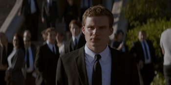 Movie still from “Ray Donovan: The Movie” (2022), directed by David Hollander – A man in a suit and tie standing in front of a group of people; Close Up shot, Over the shoulder angle
