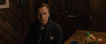 Movie still from “Raymond & Ray” (2022), directed by Rodrigo García – A man sitting at a table in front of a wooden wall; Medium shot, Over the shoulder angle