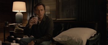 Movie still from “Raymond & Ray” (2022), directed by Rodrigo García – A man sitting on a couch holding a glass of milk; Medium shot, Over the shoulder angle