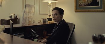 Movie still from “Raymond & Ray” (2022), directed by Rodrigo García – A woman sitting at a desk in front of a lamp; Medium shot, Over the shoulder angle