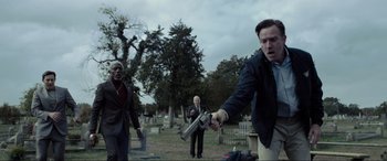 Movie still from “Raymond & Ray” (2022), directed by Rodrigo García – A man holding a gun in a cemetery with other men; Medium shot, Low angle