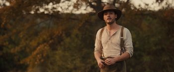 Movie still from “Redeeming Love” (2022), directed by D.J. Caruso – A man wearing a hat and suspenders standing in front of a tree; Medium shot, Over the shoulder angle