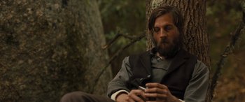 Movie still from “Redeeming Love” (2022), directed by D.J. Caruso – A man sitting on the ground using a cell phone next to a tree; Medium shot, Low angle